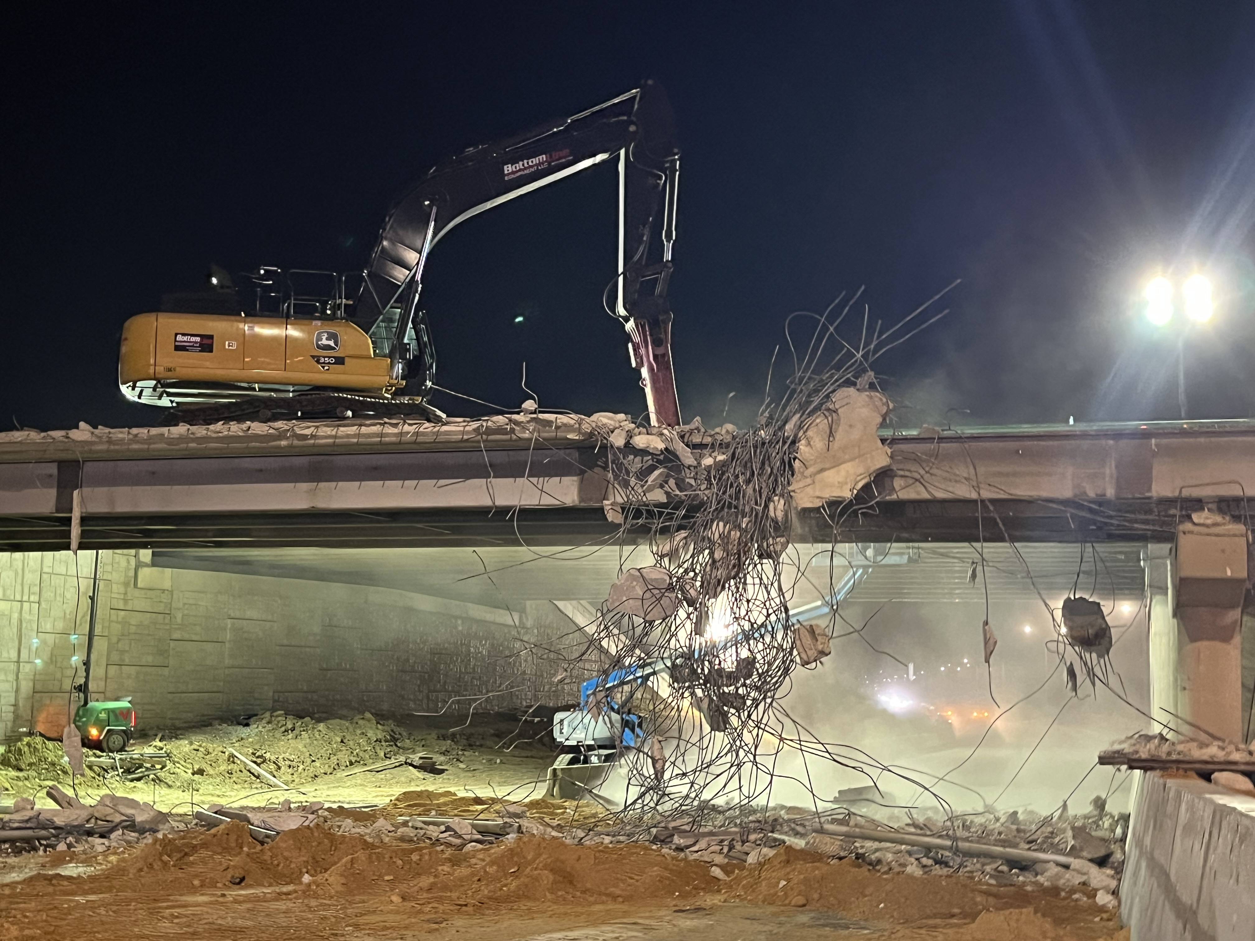 A machine with a claw atop the Lake June bridge is reaching into a tangle of metal wires that are coming out of the bridge in the middle of the construction site at night. 
