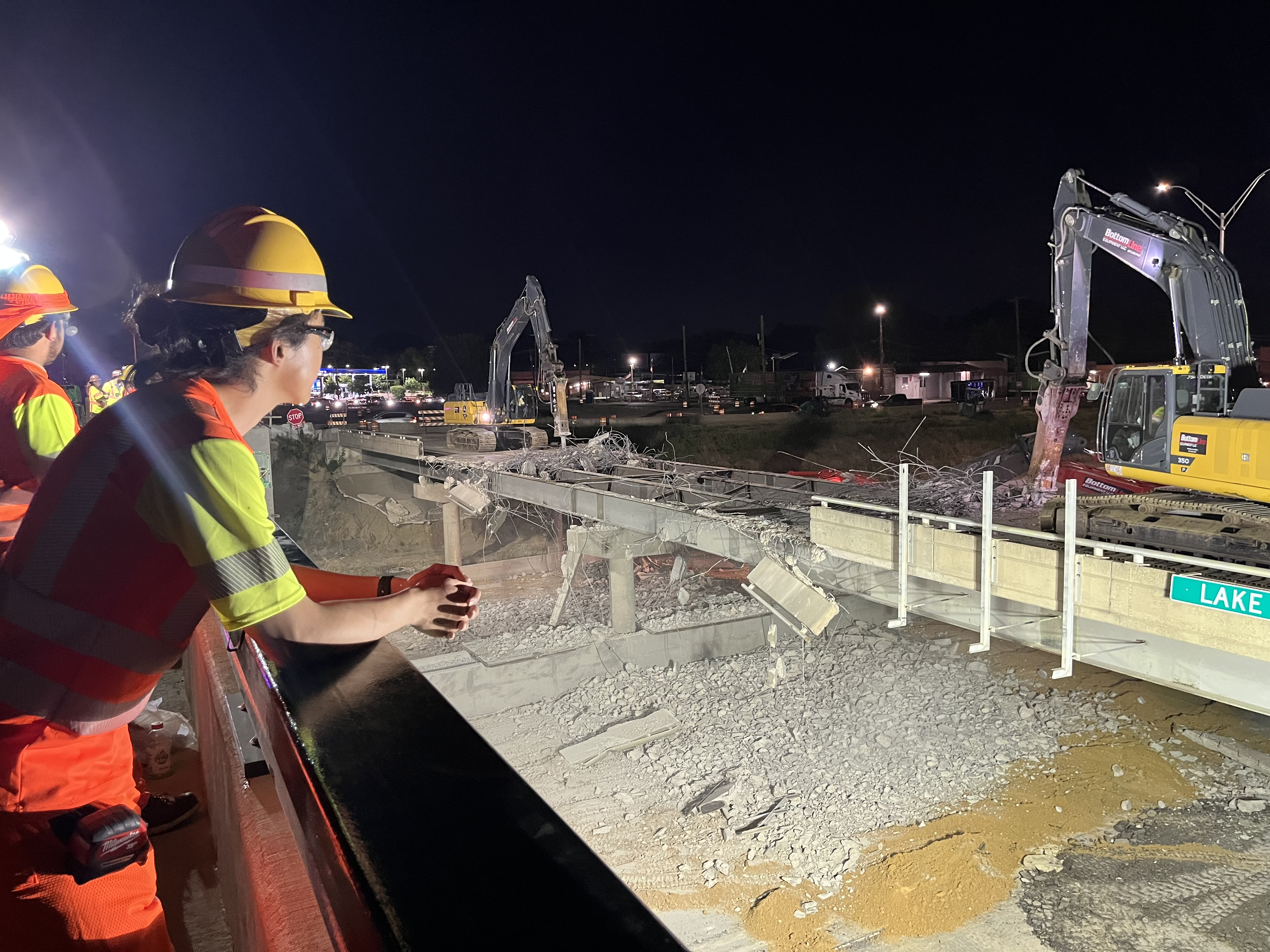 A worker in an orange vest and hard hat safety PPE is leaning on their elbows on the railing of one bridge looking over at the construction occuring across from them on the Lake June bridge.
