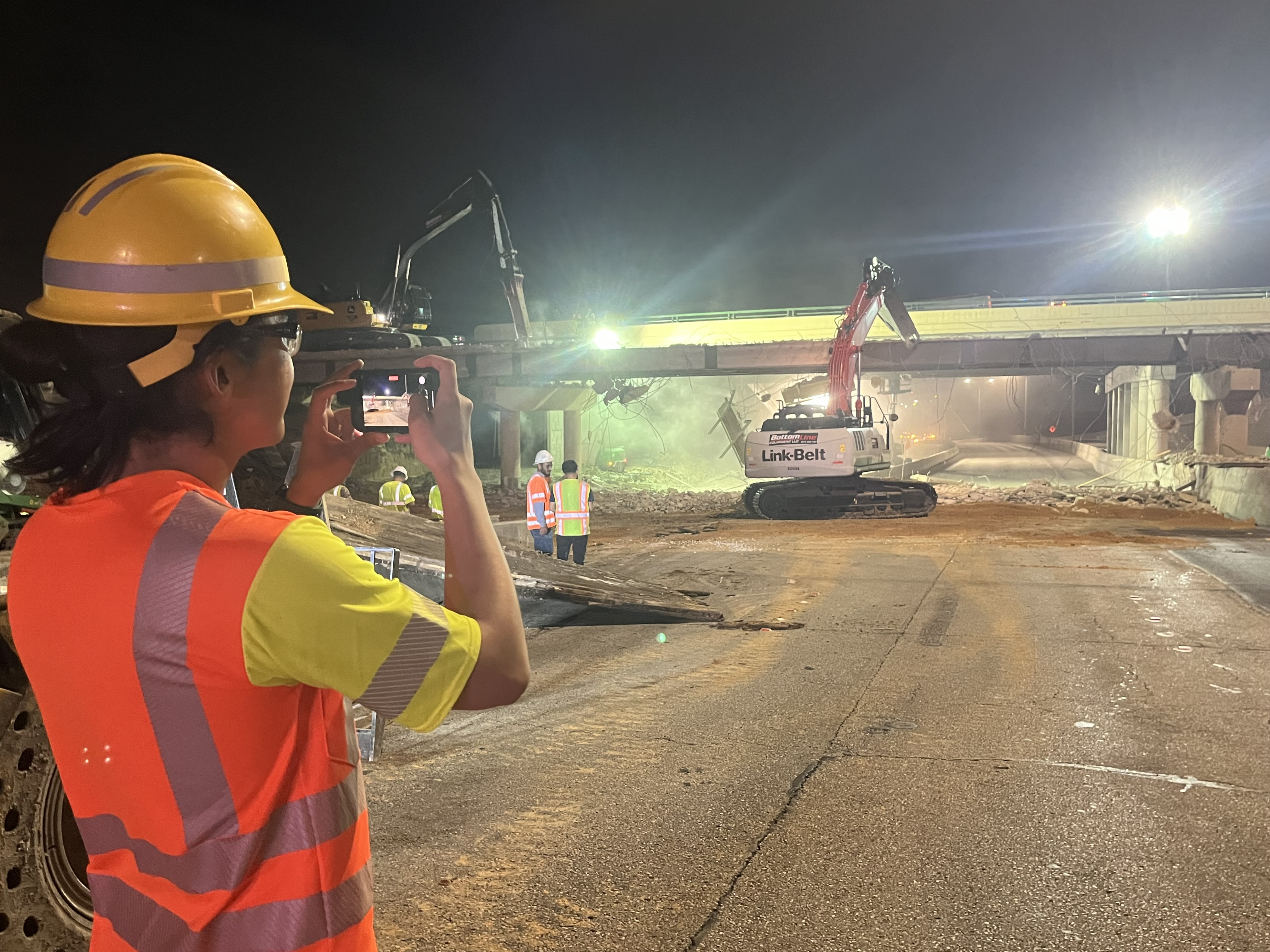A worker in an orange PPE vest and hard hat is taking a picture of the construction site with his phone. In the construction site is the bridge, a link belt machine, and several other workers. 