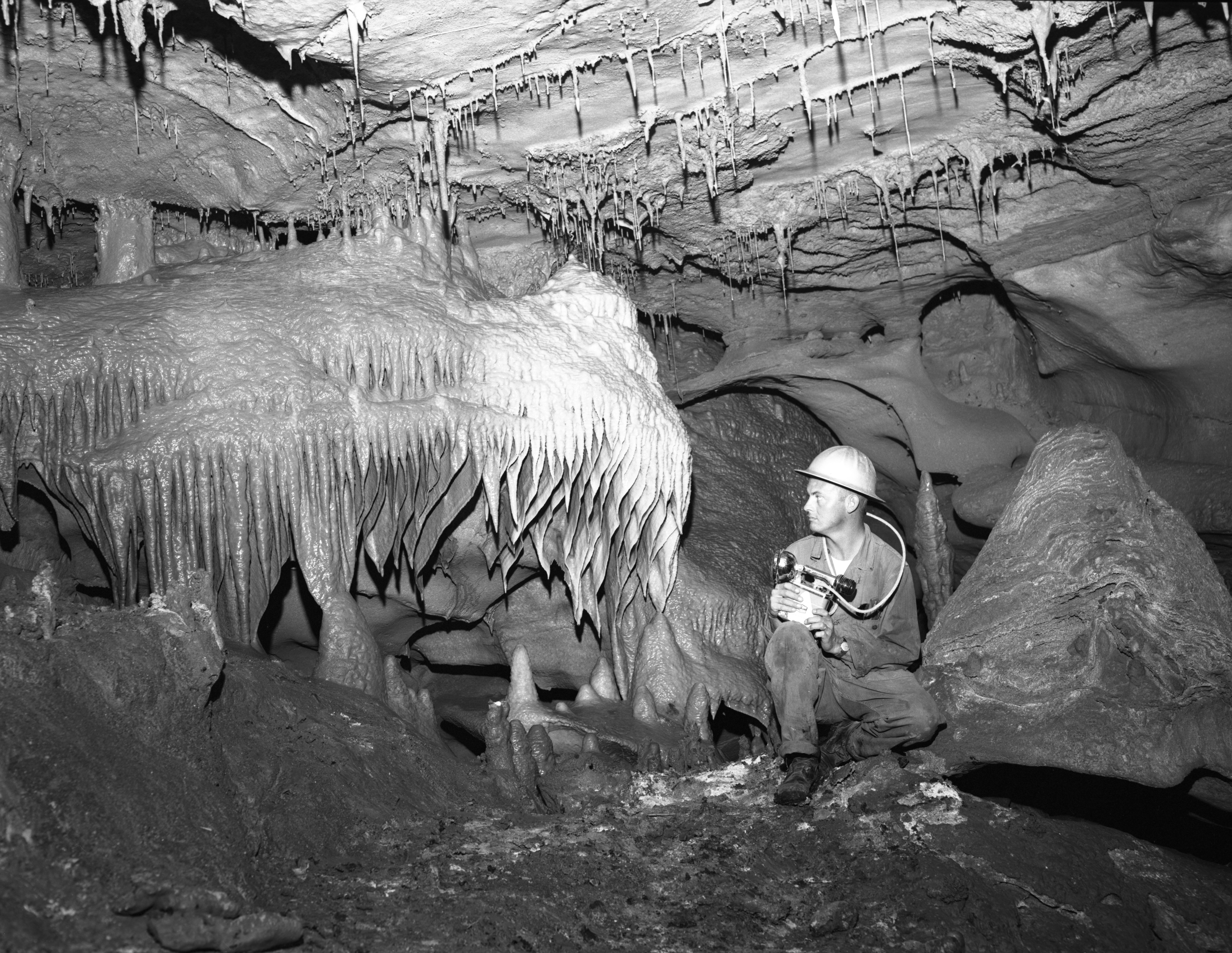 An old black and white photo of a worker holding some sort of equipment and wearing a hard hat inside inner space cavern, crouched beside a strange rock formation that looks like it's melting.