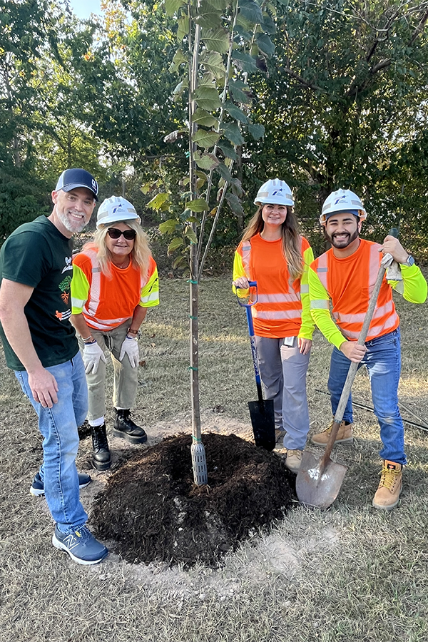 Workers in safety gear pose with shovels around a recently planted tree.