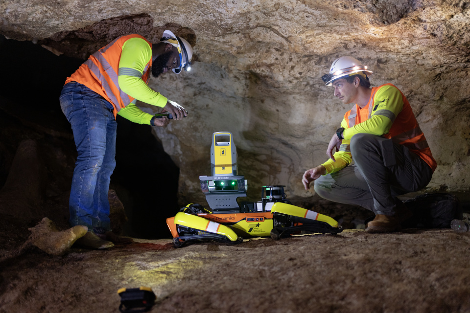 A photo of two men in orange safety vests and hard hats crouched beside a four legged walking robot on a stone ledge inside Inner Space Cavern. One man is holding what appears to be a remote operating device. 