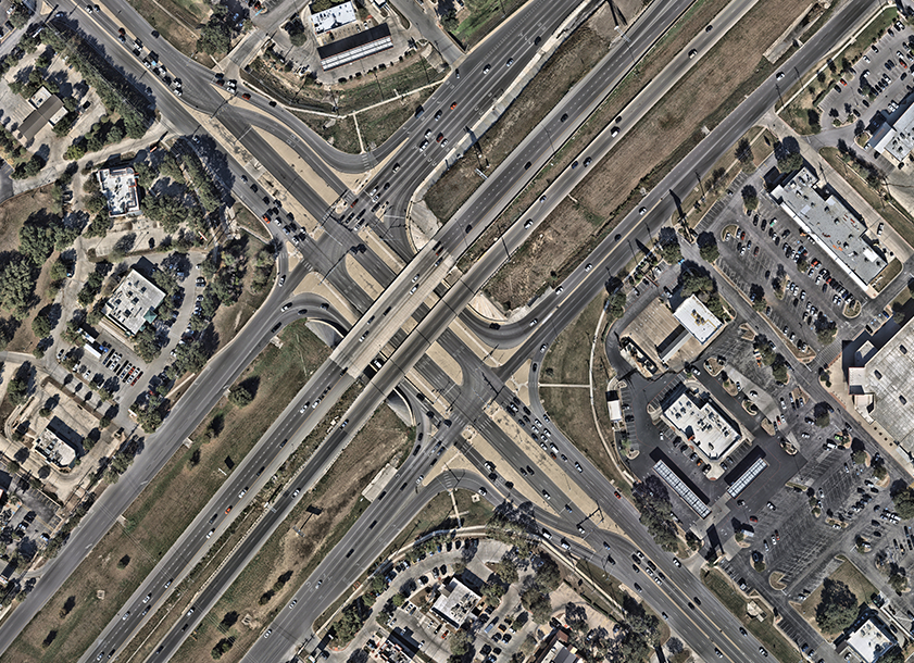 Aerial view of a displaced left-turn (DLT) intersection with an overpass, showing left-turn traffic rerouted across opposing lanes at signalized crossovers before the main intersection, enabling simultaneous left-turn and through movements.
