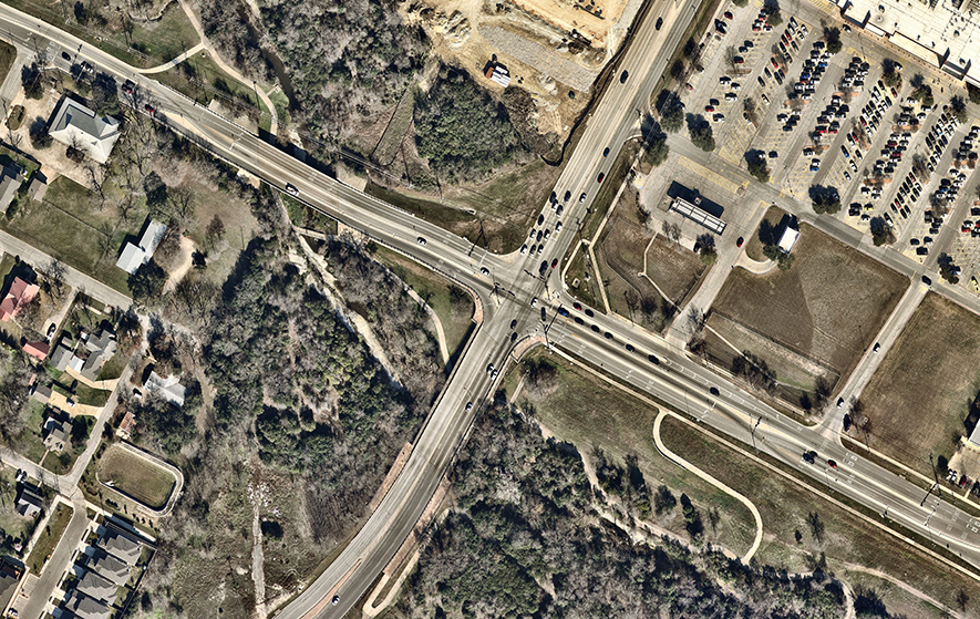 Aerial view of a displaced left-turn (DLT) intersection showing left-turn vehicles crossing opposing traffic at upstream signalized crossovers before reaching the main intersection, enabling simultaneous through and left-turn movements across the intersection.