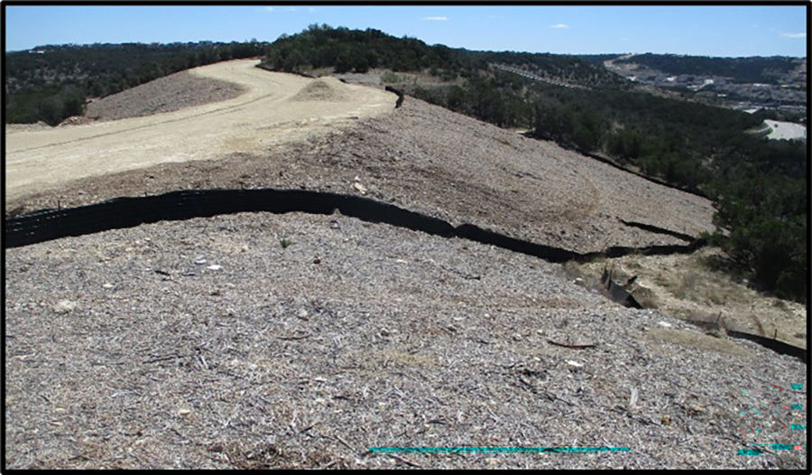 A hillside slope covered with mulch for stabilization. There is also sediment control fence installed along the contour of the hillside. There is a dirt road on top of the hill and there is sparse vegetation in the background.
