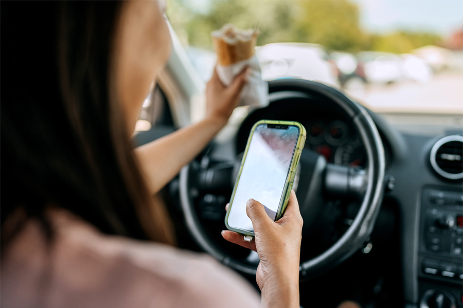 Over the shoulder view of woman text messaging while driving, she is holding sandwich in other hand.