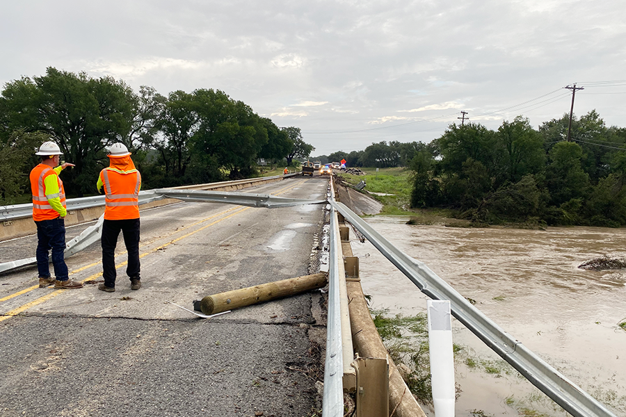 TxDOT roadway crew in orange vests stand on a bridge damaged by flood waters.